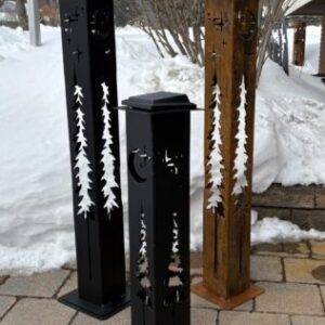 Three tall solar garden light columns with evergreen tree cutouts plus star and crescent moon details, displayed on a stone walkway beside snowbanks; two are black and one has a rust patina finish.